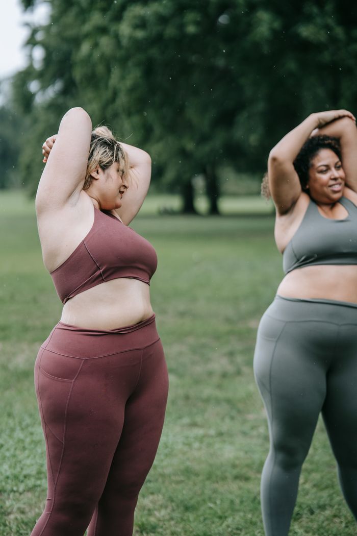 services-02 Two women in activewear stretch in a park, promoting fitness and body positivity.