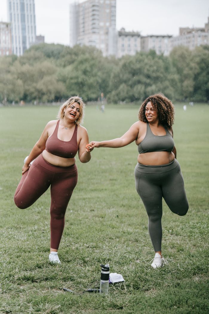 heros-img Two women stretching together in a park, smiling and enjoying their workout.
