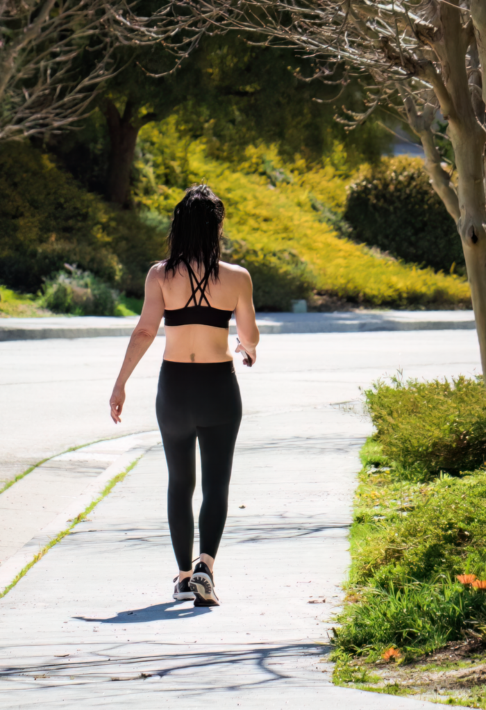 services-03 A woman exercises while strolling on a city sidewalk in activewear on a bright, sunny day.